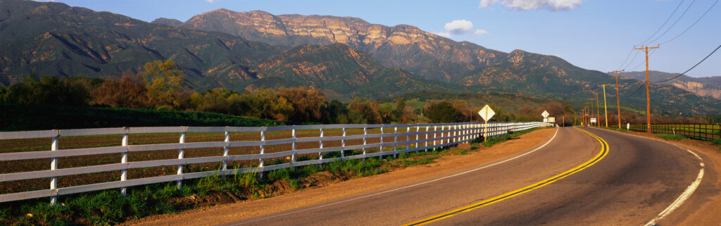 This is scenic Route 150 with the Topa Topa Mountains in the background. The road curves and there is a white fence along the left hand side of the road marking the nearby ranch property. This area is known as upper Ojai.