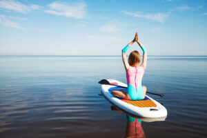 Woman doing yoga on sup board with paddle. Mediatative pose, back view - concept of harmony with the nature, free and healthy living, freelance, remote business.